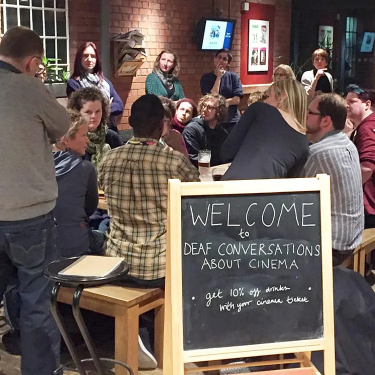 Group of people gathered in a brick-walled venue for a "Deaf Conversations About Cinema" event, with a welcome sign visible.