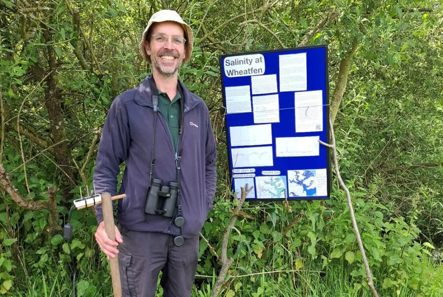 Man holding tools and binoculars standing beside an educational display board about salinity in dense vegetation.