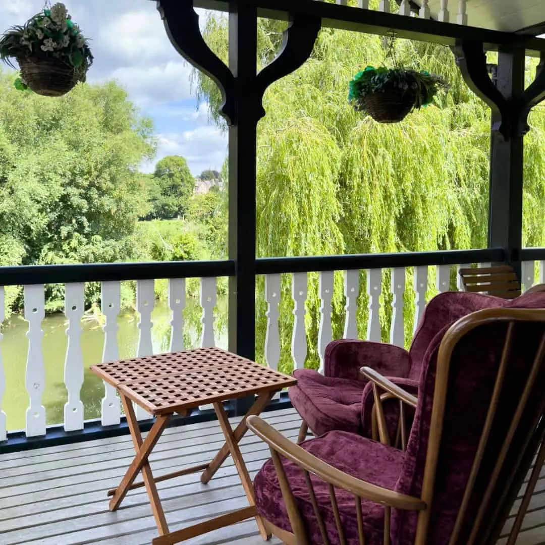 Covered riverside deck with burgundy armchairs, small table, and hanging baskets overlooking weeping willow trees and water.