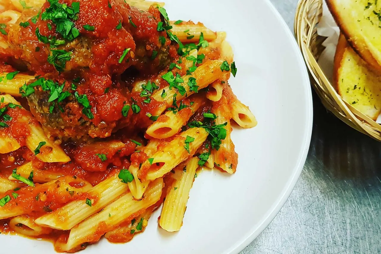 Penne pasta in tomato sauce with fresh parsley garnish, served on a white plate with bread basket alongside.