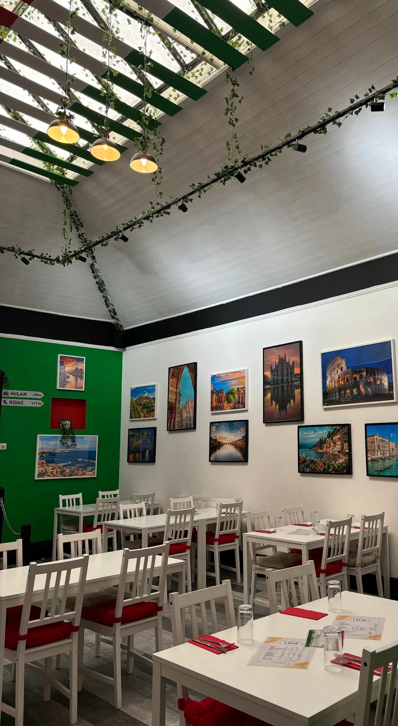 Modern restaurant interior with white tables and chairs, green and cream walls decorated with European landmark photographs, and a green pergola ceiling with hanging pendant lights.