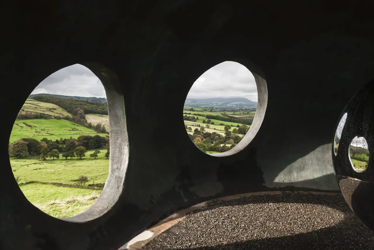 Looking out through circular openings in a stone structure towards rolling green hills and patchwork fields.