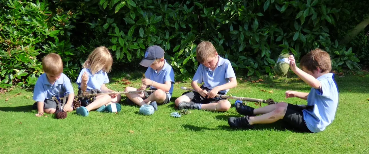 Children in blue school uniforms sit on grass examining natural objects during an outdoor educational activity.