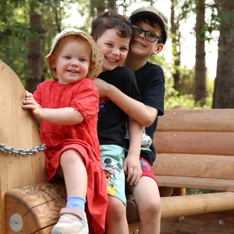 Three children smiling together on wooden playground equipment in a forest setting.