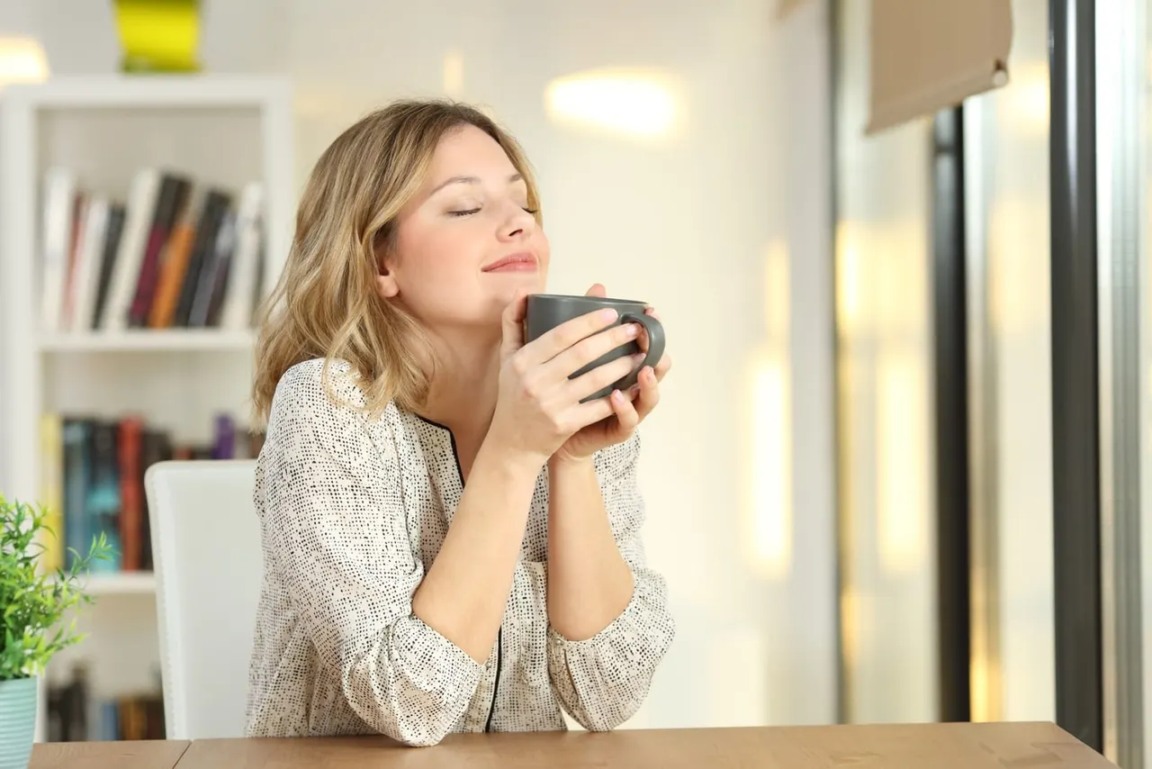 Woman holding a dark mug while relaxing indoors, with bookshelves visible in the background.