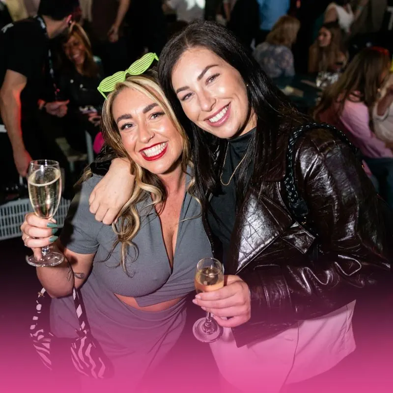 Two women holding champagne glasses at a busy indoor event, smiling at the camera with a crowd visible behind them.