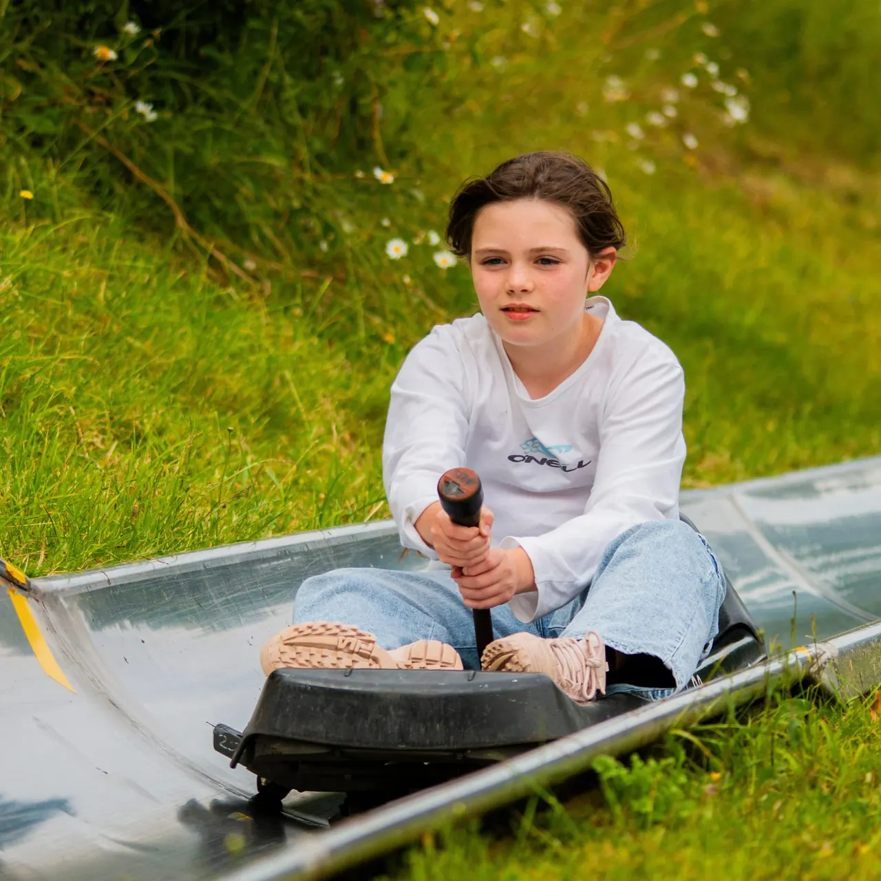Child in a bobsleigh on an indoor ice track, holding the push handle and wearing casual clothing.