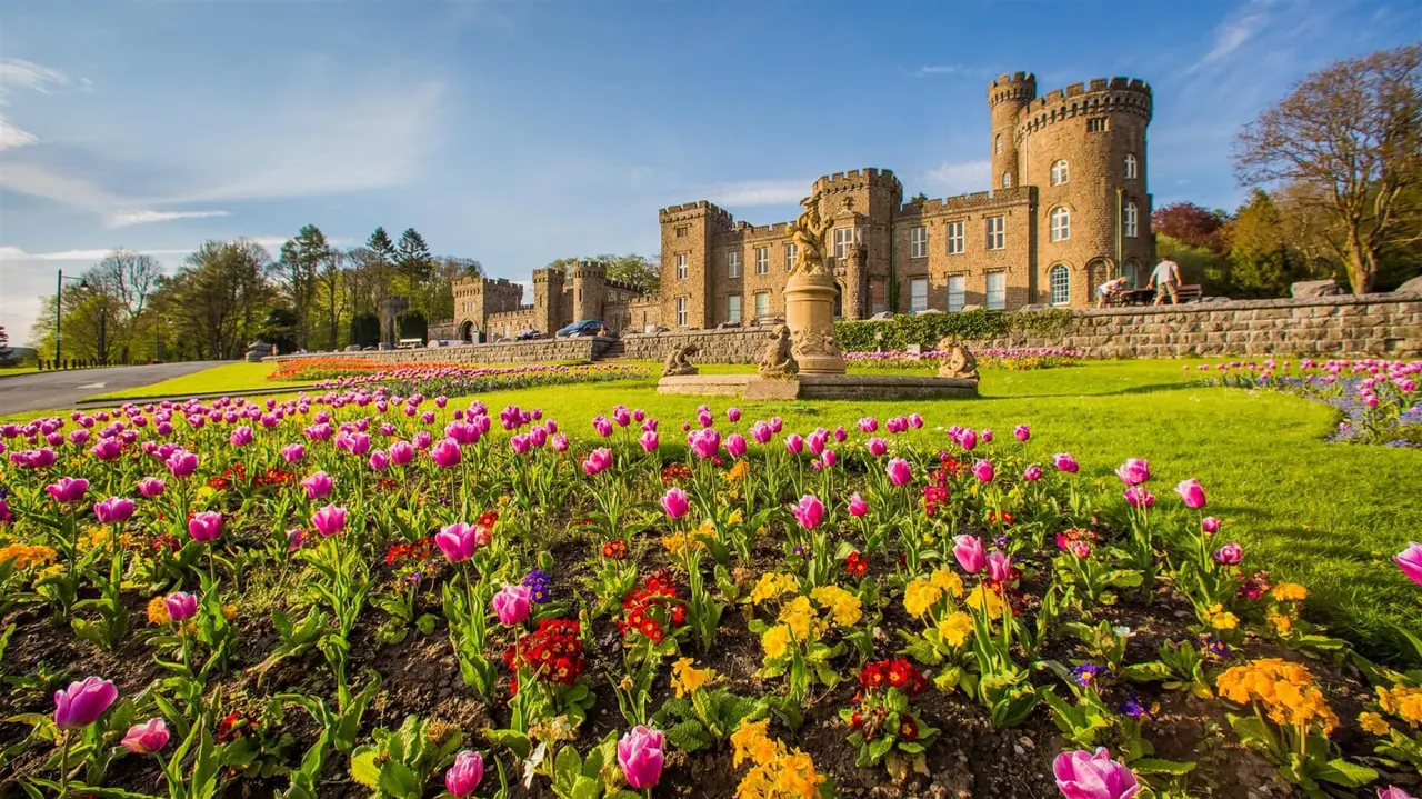 Spring flower beds with pink tulips and colourful blooms in front of a stone castle with crenellated towers.