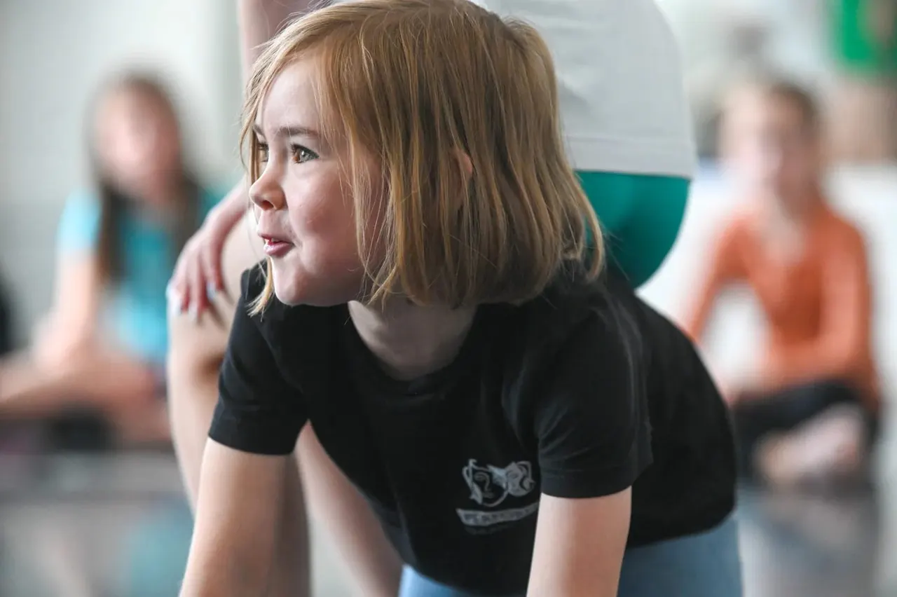 Child in black t-shirt participating in a drama or movement activity with other young people blurred in the background.