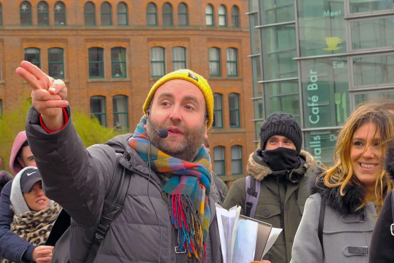 A tour guide wearing a yellow beanie and microphone headset points while speaking to a group of visitors on an urban street, with Victorian brick buildings behind them.