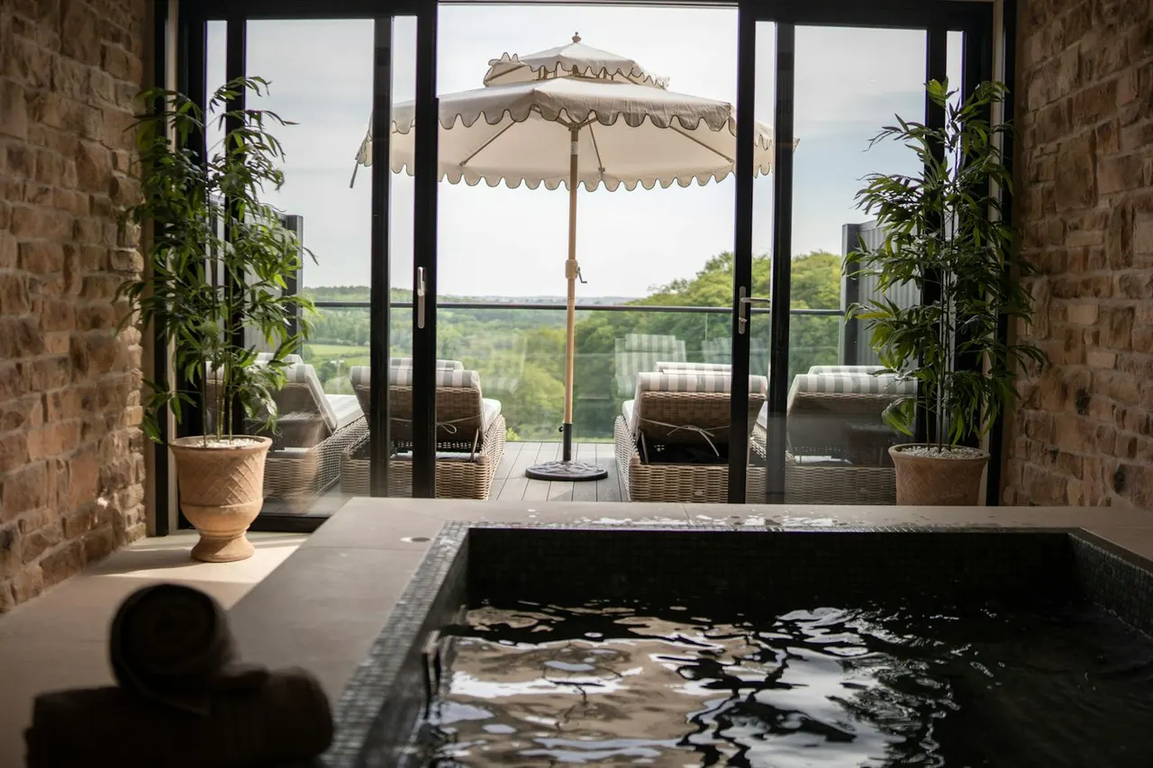 Indoor spa pool with countryside views through large glass doors opening to a terrace with parasol and loungers.