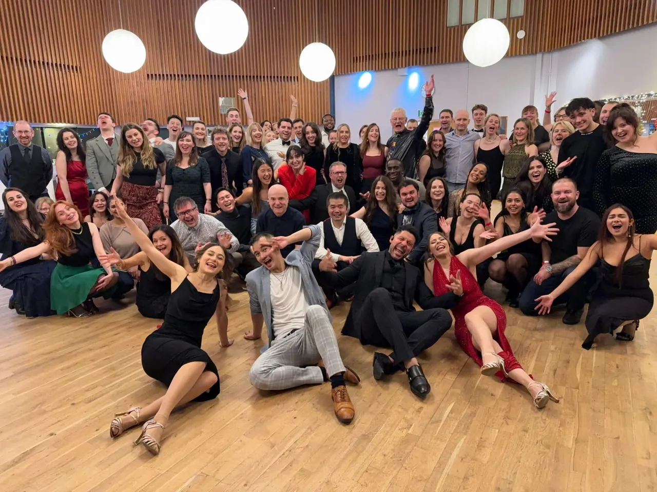 Large group of people posing together on a wooden dance floor in a modern hall with circular lights overhead.
