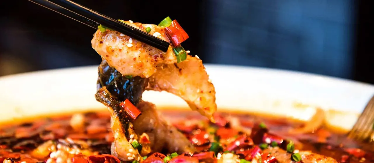 Chopsticks lifting a piece of cooked prawn and vegetable from a spicy Asian dish on a white plate.