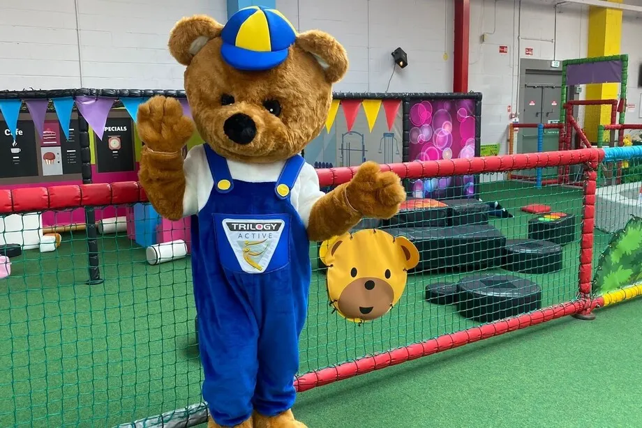 Large mascot character wearing blue dungarees and cap standing in soft play arena with climbing equipment and padded structures visible behind.