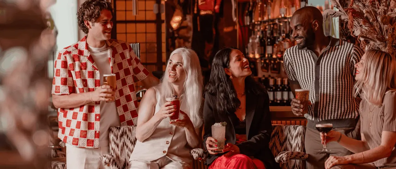 A group of six people socialising and smiling at a bar, holding drinks with warm amber lighting and bottle shelves visible behind them.