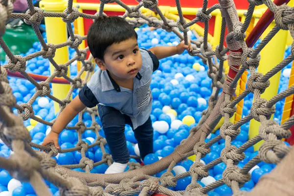 Young child climbing through a rope net obstacle course above a ball pit.