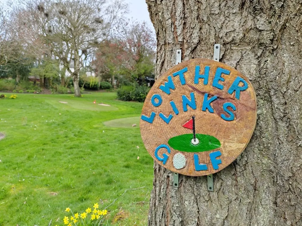 A wooden sign reading "Mother Link's Golf" with a painted hole flag is mounted on a tree trunk overlooking a grass pitch and putt course.
