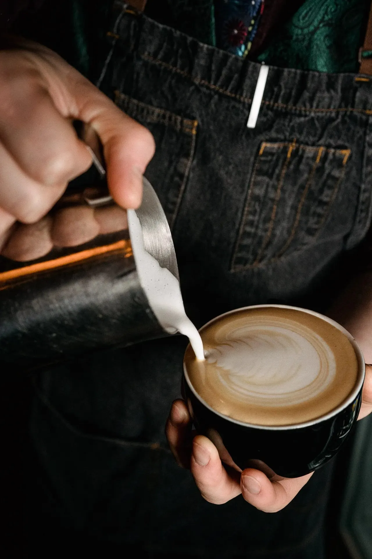 Barista pouring steamed milk into a black cup of coffee to create latte art.