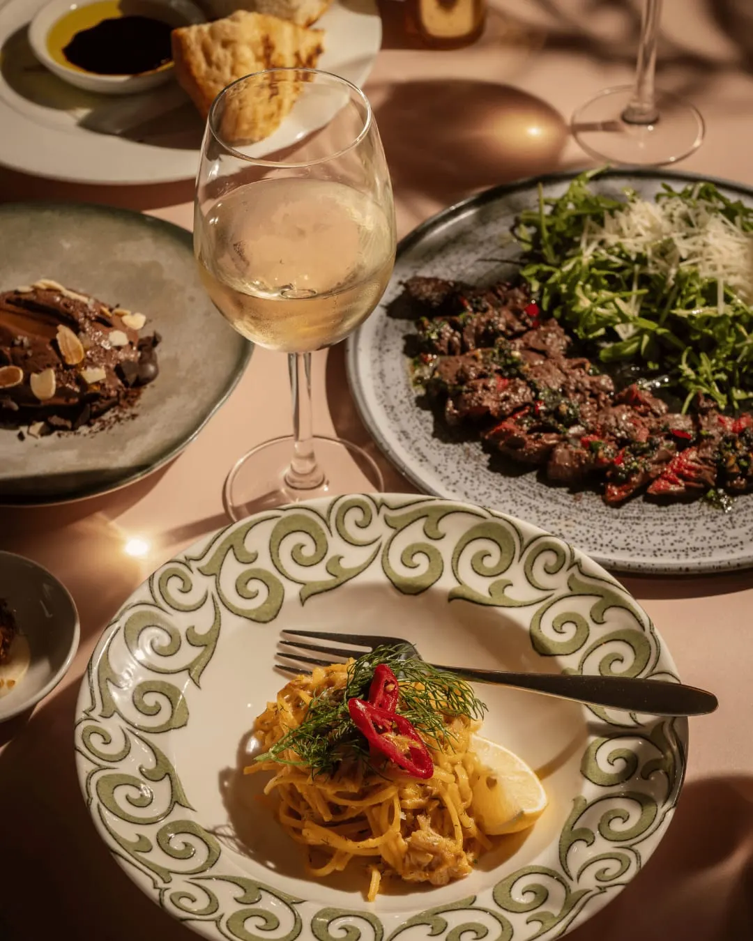 Fine dining table set with pasta dish, wine glass, and grilled meat plates with salad and garnishes.