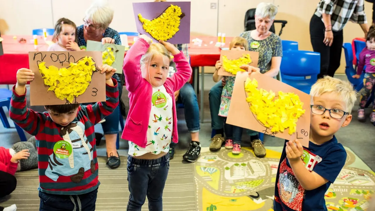 Young children display yellow chicken artwork at an intergenerational craft activity with adults supervising.