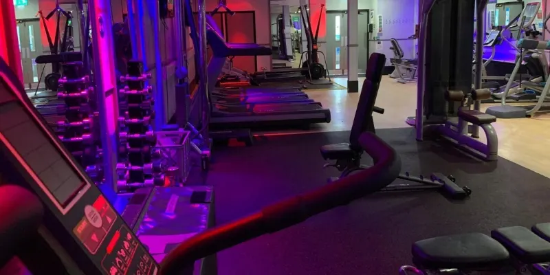 Modern gym interior with weight machines and cardio equipment illuminated by neon pink and blue lighting.