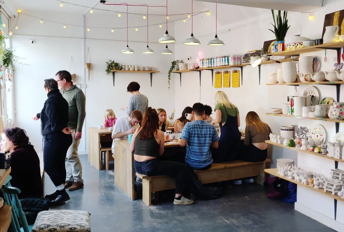 Groups of adults and children seated and standing in a bright workshop café with wooden shelving, pendant lights and potted plants.