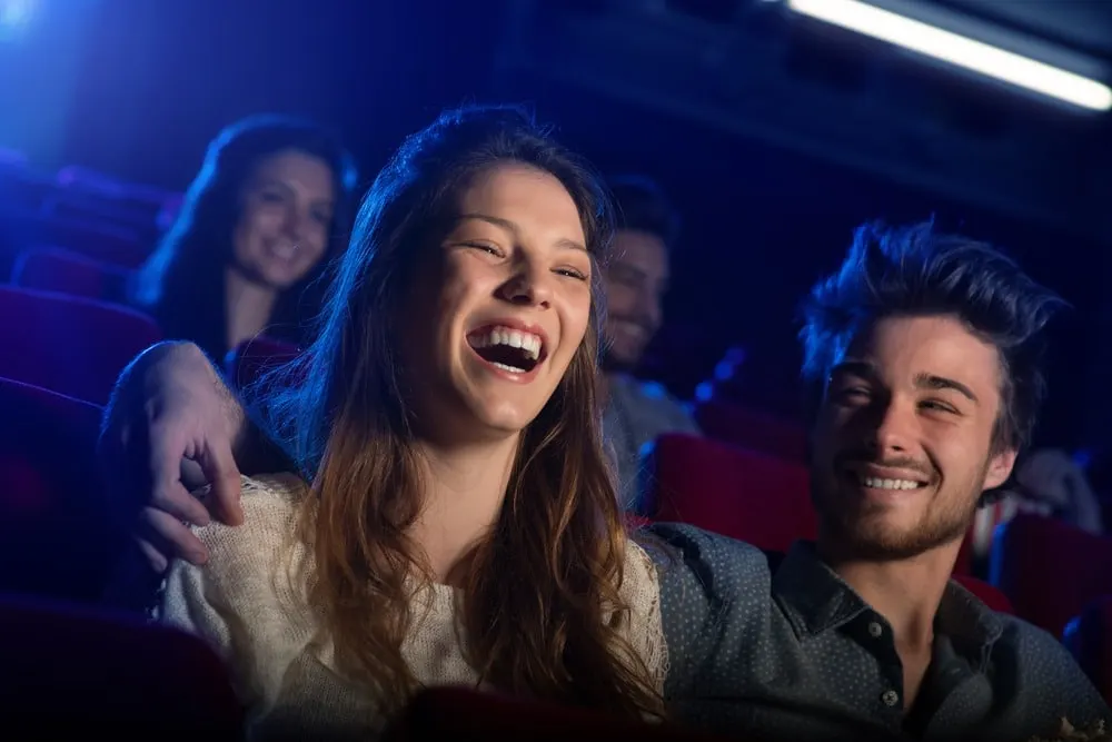 Young couple laughing together in a cinema auditorium with other patrons visible in the background.