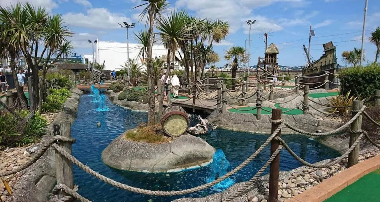 A themed mini-golf course with water features, palm trees, rope barriers and pirate-themed structures including a wooden tower and ship wreckage.