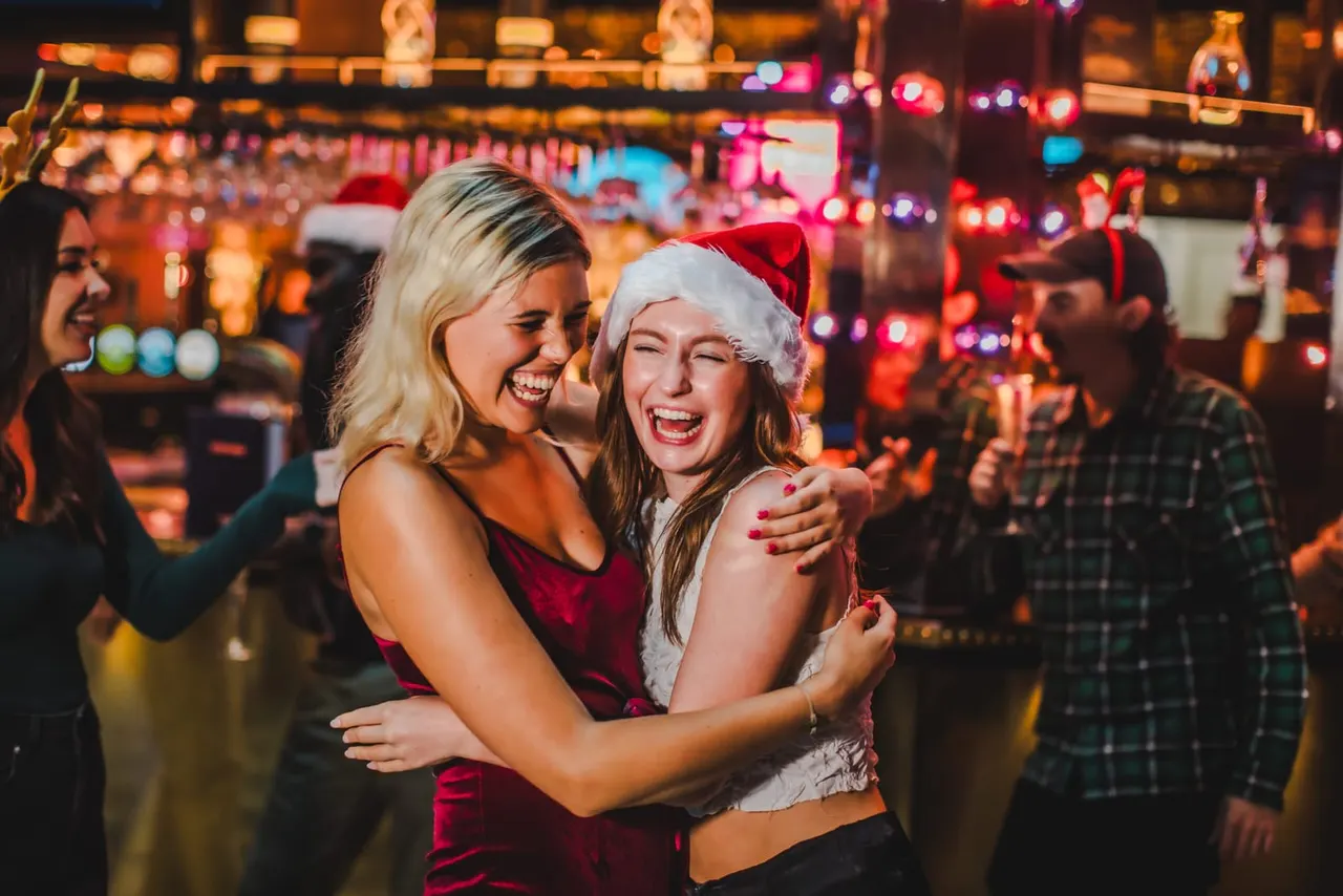 Two young women embracing and laughing at a festive party in a brightly lit venue with colourful neon lights and other guests in the background.