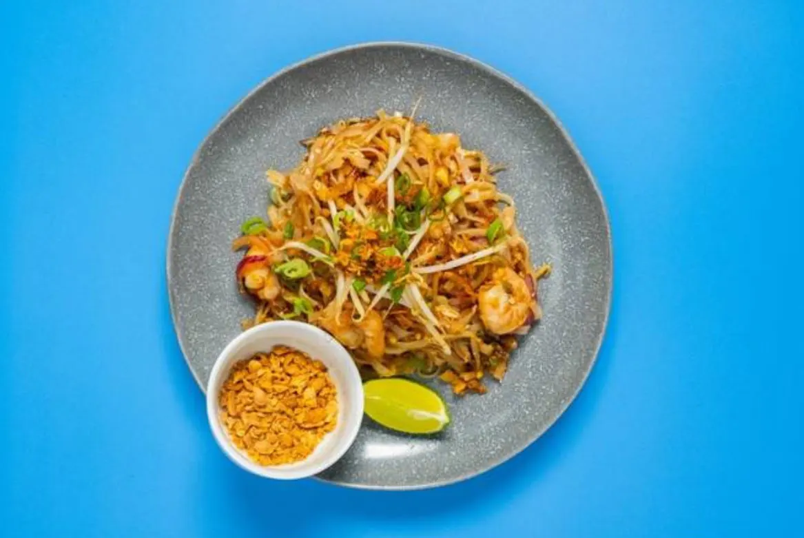 Stir-fried noodles with prawns, green onions and chilli, served with peanut sauce on a grey plate against blue background.