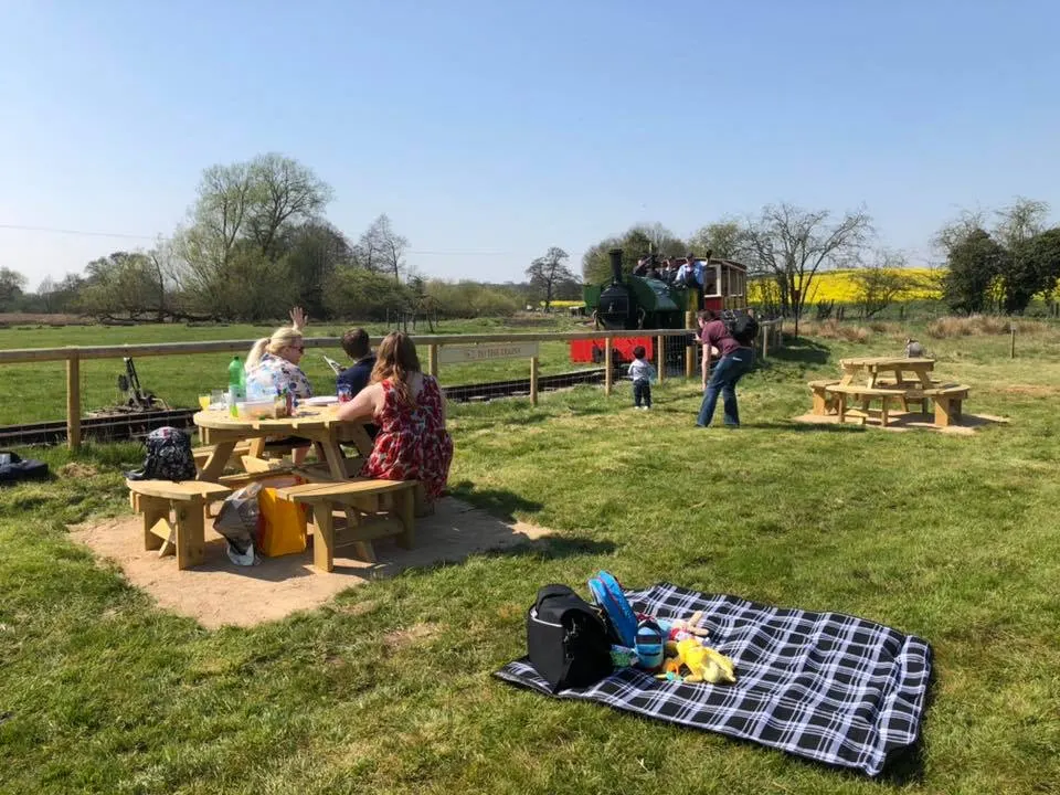 Families picnicking and children playing at outdoor wooden tables in a grassy field beside a heritage railway line.
