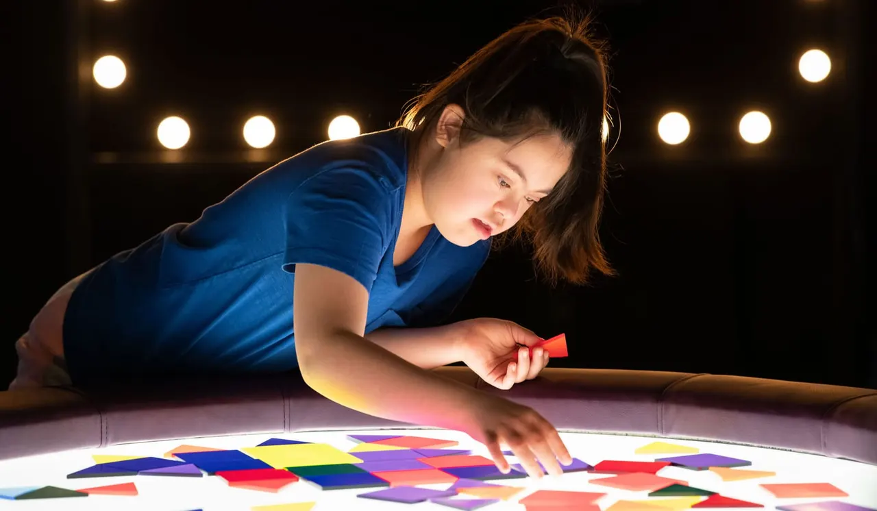 Young child in blue shirt leaning over illuminated interactive display with colourful tiles, holding stylus, surrounded by stage lights.
