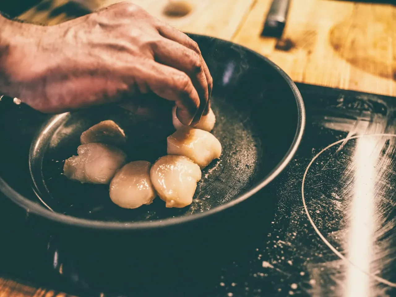 A person's hand places peeled potatoes into a dark cooking pan on a wooden work surface.