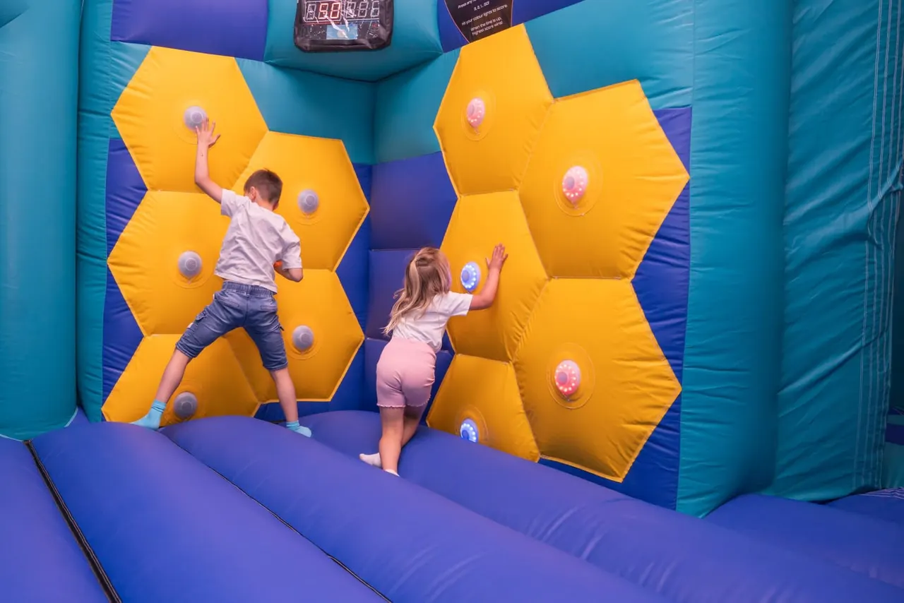 Two children playing on a colourful inflatable activity wall with glowing target buttons in an indoor play centre.