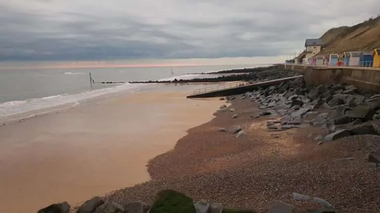 Sandy beach with shingle foreshore and colourful beach huts on clifftop under cloudy sky.