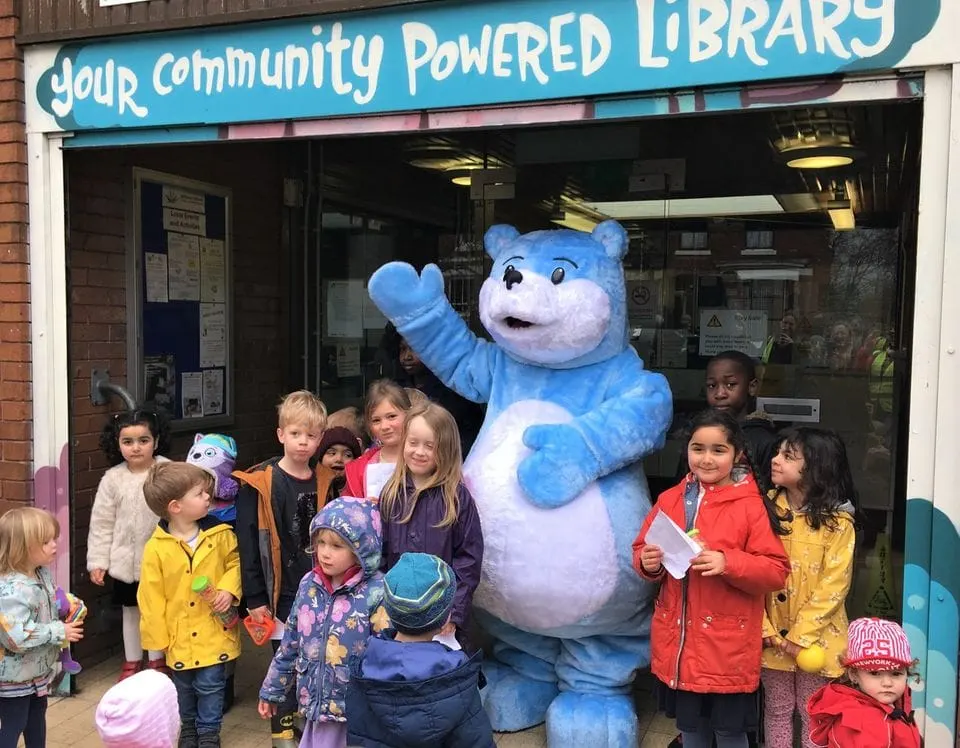 A group of children posing with a large blue mascot costume outside a community library entrance.