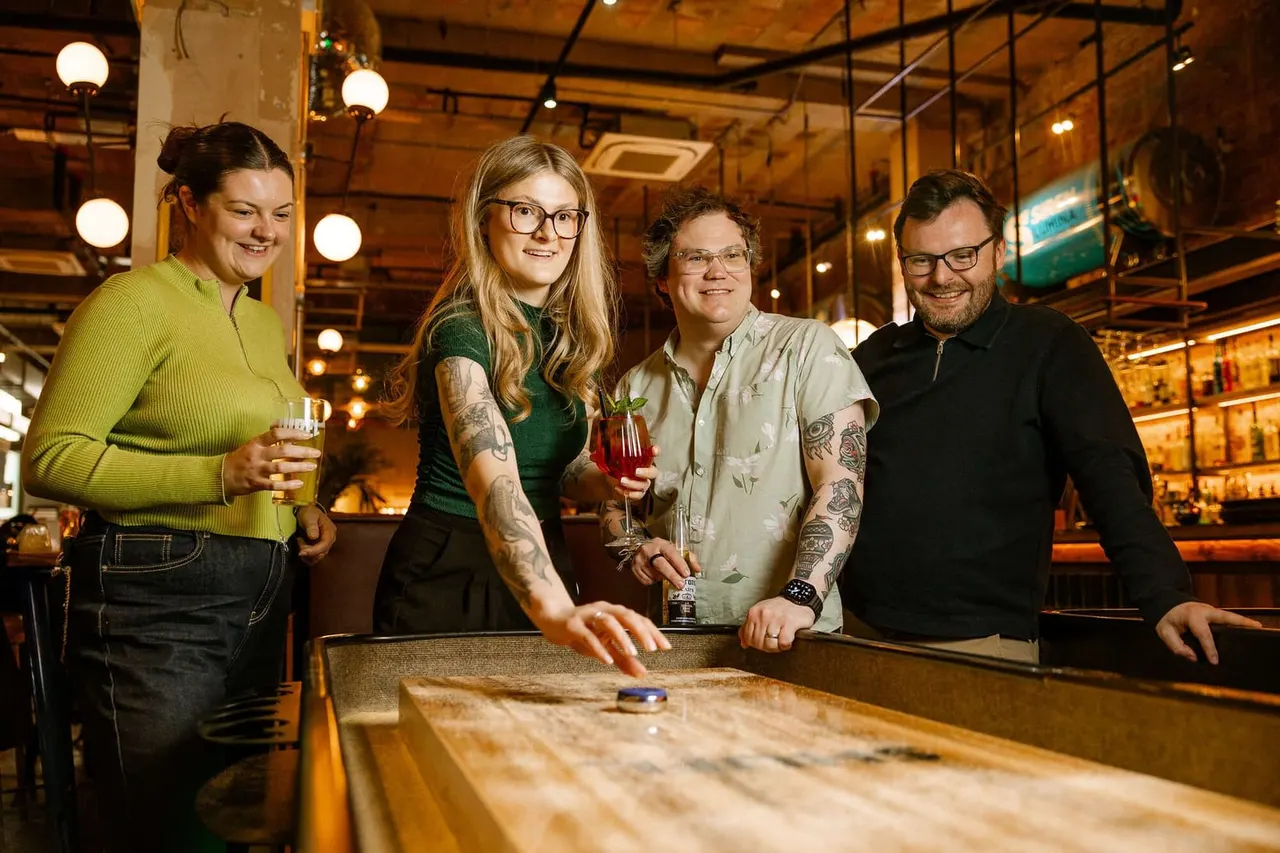 Four people smiling at a shuffleboard table in a warm-lit bar with drinks in hand.