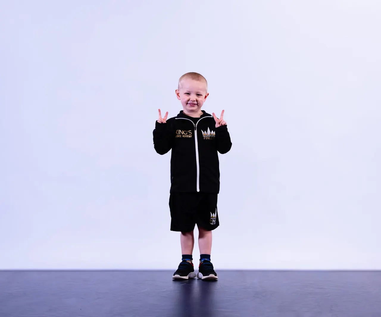 Young child in black dance academy outfit making peace signs on stage against light blue background.