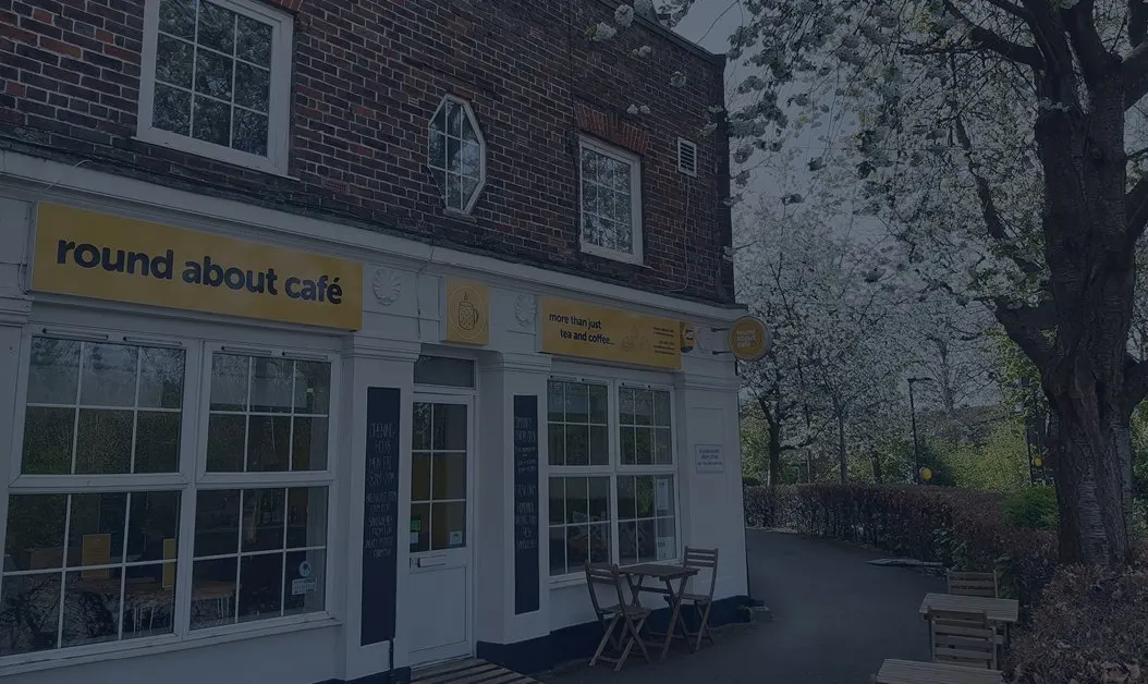 Exterior of Round About Café with white-framed windows, yellow signage, and outdoor seating beside a large ivy-covered tree.