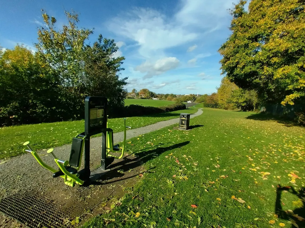 Outdoor fitness equipment in a park with a curved pathway, green fields, and tree-lined countryside views under blue sky.