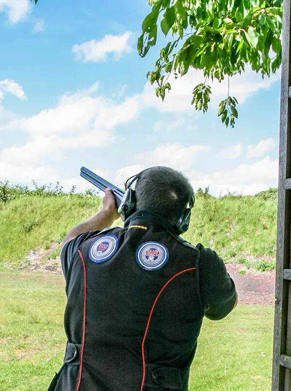 Clay pigeon shooter aiming at target with shotgun at outdoor range under clear sky.