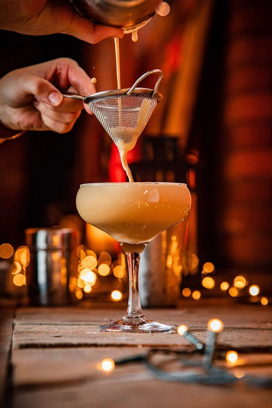 Bartender straining a cocktail through a fine mesh sieve into a coupe glass, surrounded by candlelight.