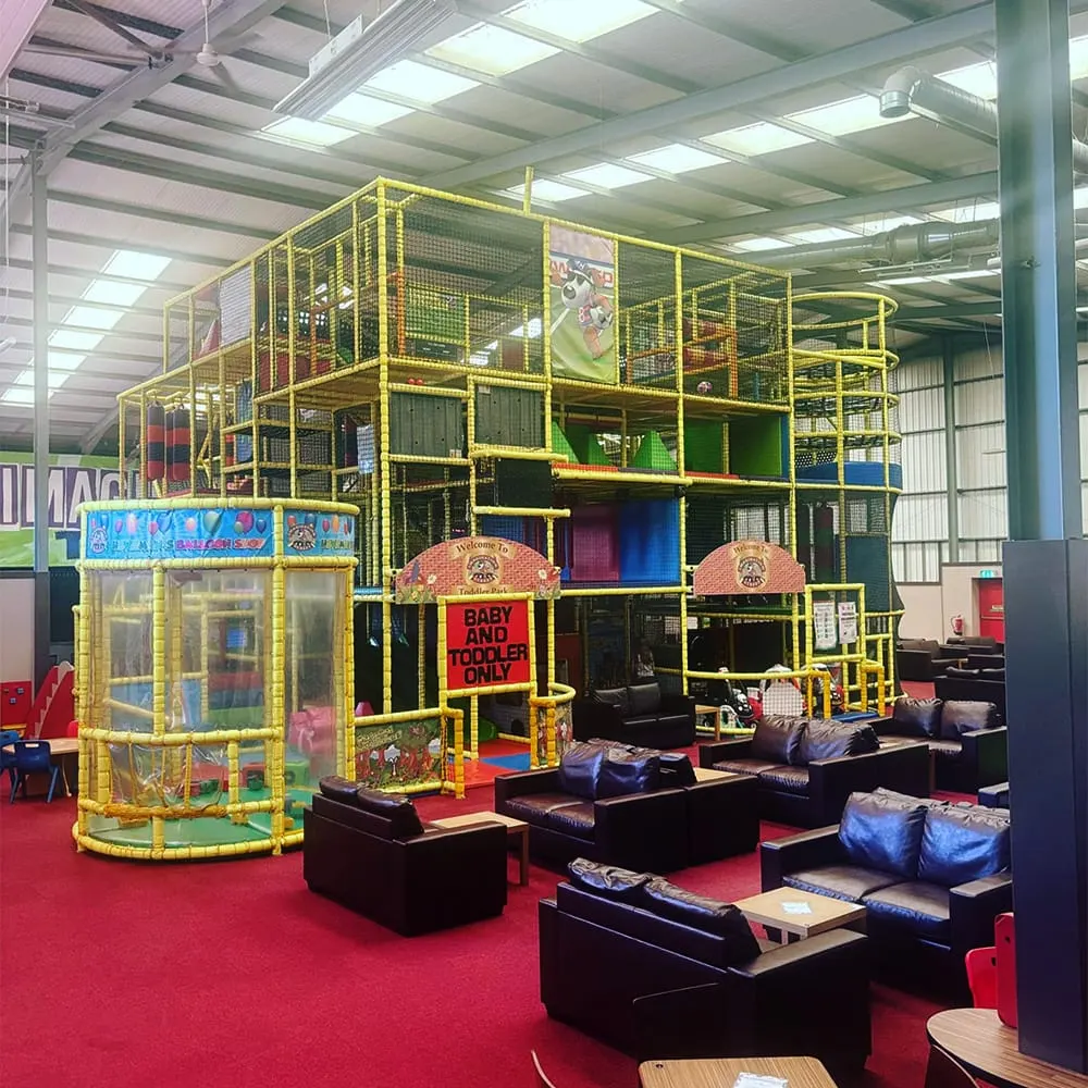 Indoor play area with large yellow metal climbing structure, ball pits, and soft play equipment on red flooring.