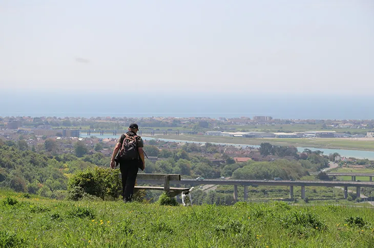 Hiker with backpack stands at wooden bench on grassy hillside overlooking countryside landscape and distant river.