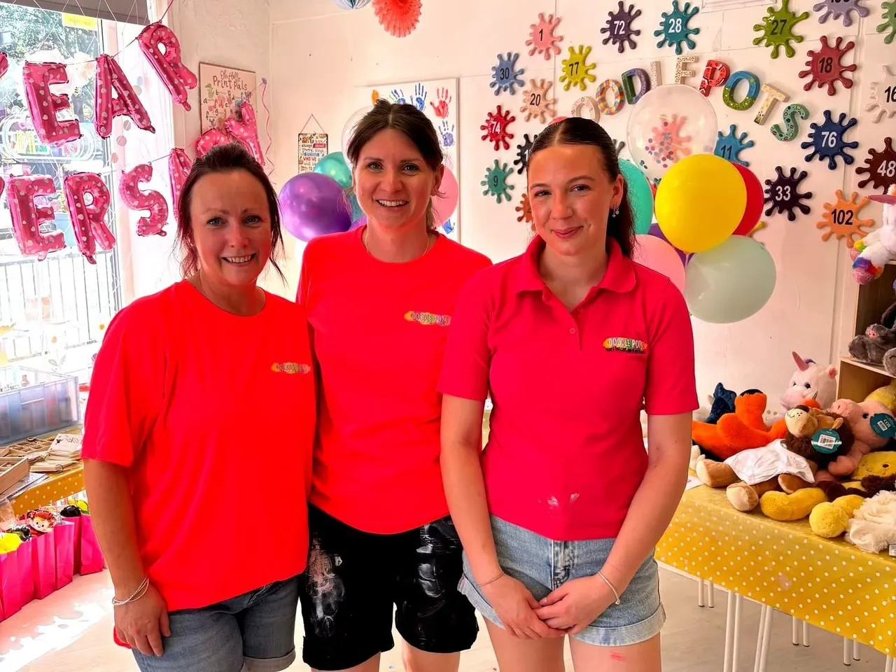 Three staff members in red uniforms smile at the camera in a colourful children's craft studio decorated with balloons, handprints and numbered virus decorations.