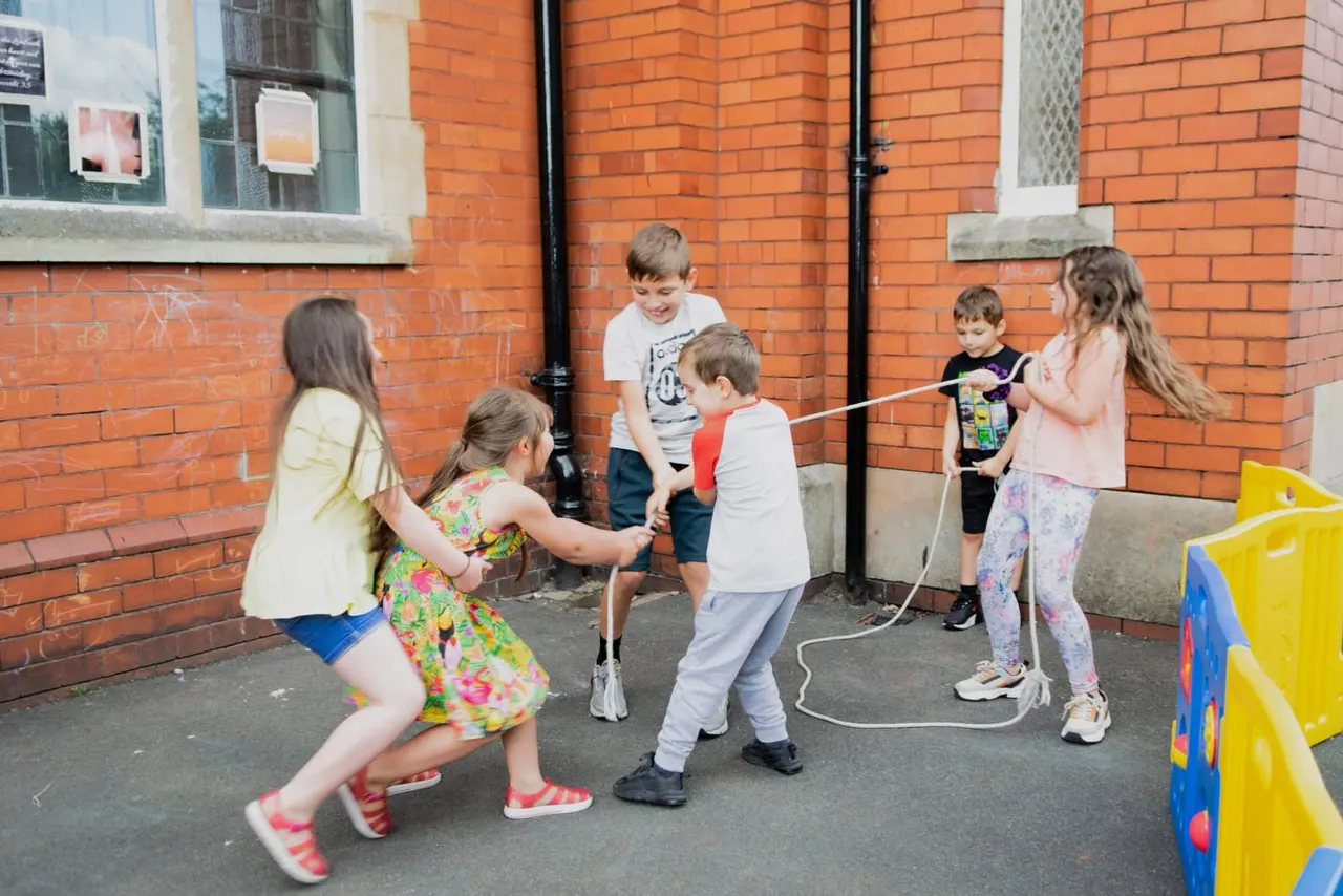 Group of children playing with a skipping rope in a paved outdoor courtyard against a red brick building.
