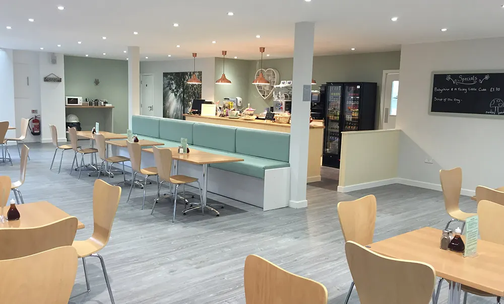 Modern café interior with wooden tables and chairs, service counter with turquoise seating, and vending machines in background.