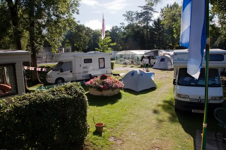 Campsite with caravans, tents and motorhomes parked on grassy pitches amongst trees and flowering shrubs.