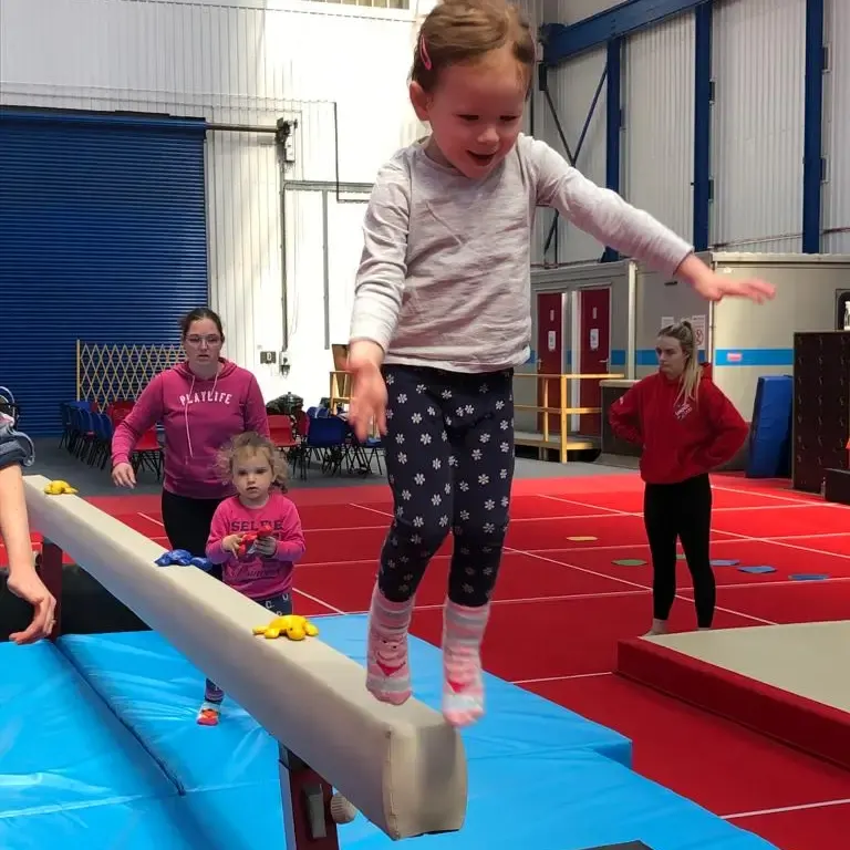 A young child balances on a low beam during a gymnastics session at an indoor facility, with instructors and other children watching nearby.