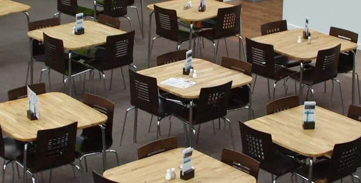 Multiple wooden tables with black chairs arranged in a dining or event space, each set with water bottles and condiments.